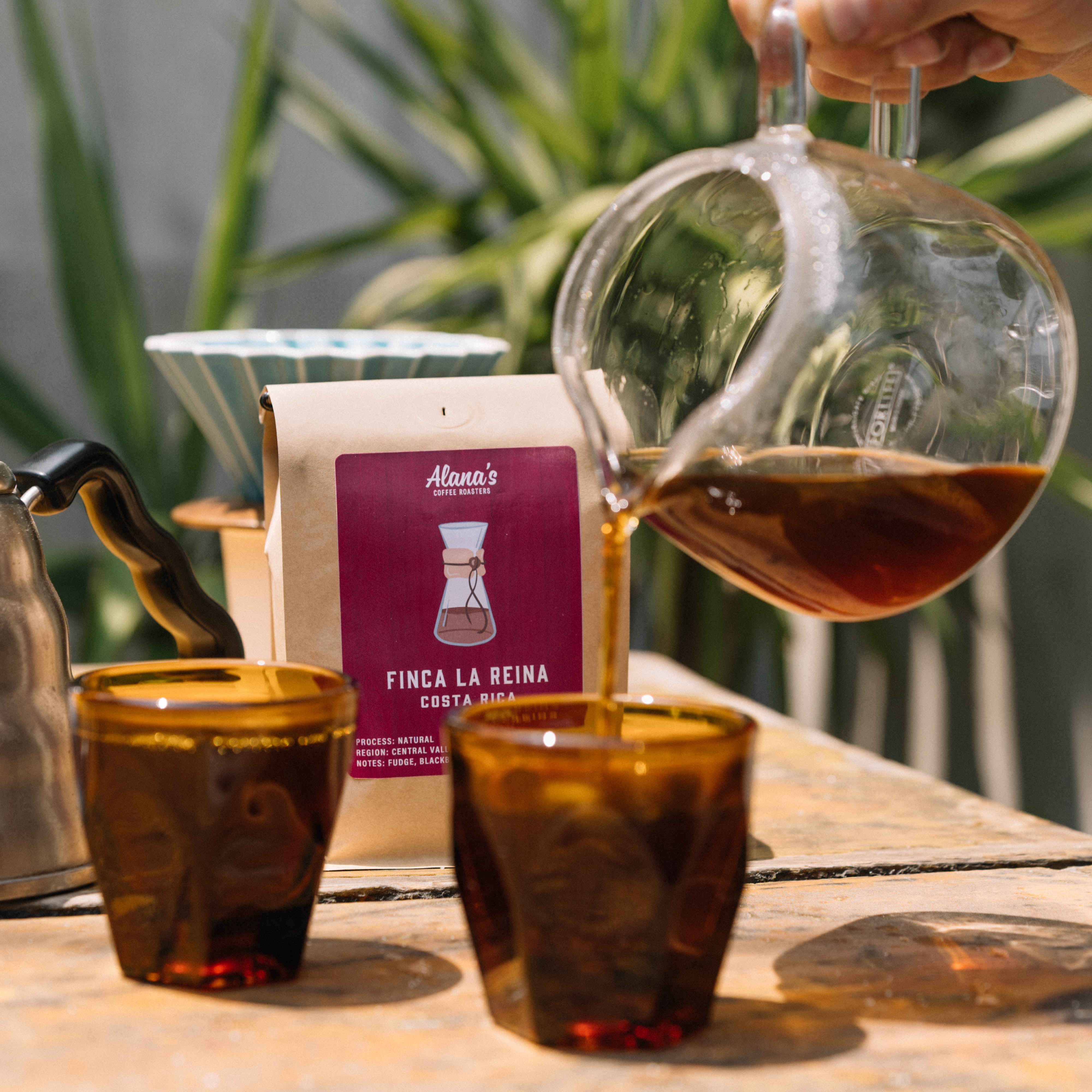Person pouring coffee from a glass carafe into two small cups on a wooden table with a coffee bag in the background.