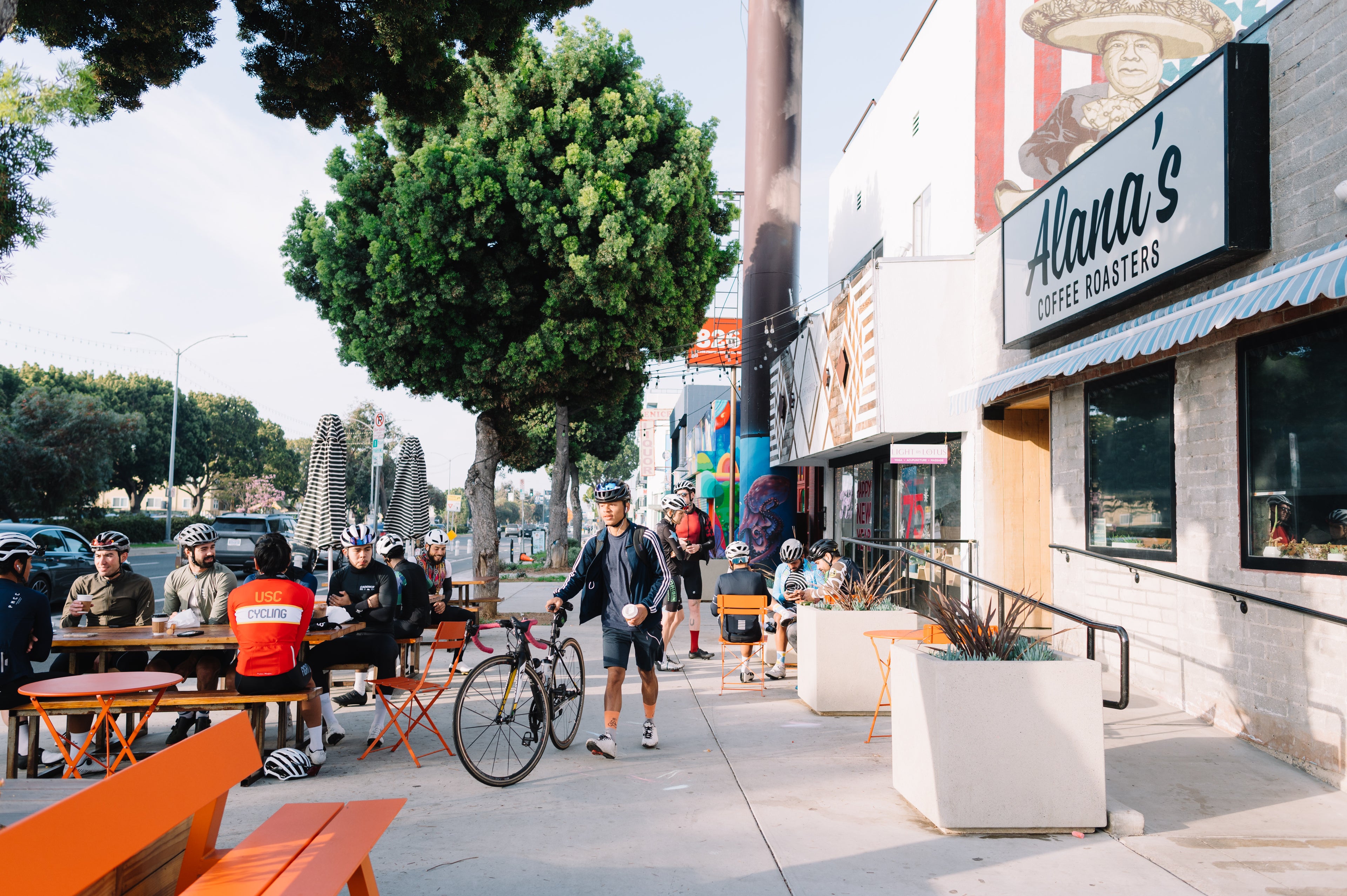 The sidewalk outside Alana's in Mar Vista, California. A group of bikers sit on bright orange patio furniture across the walkway from the front door of Alana's.