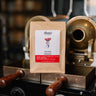 Close-up of a brown coffee bag labeled Inkingi Rwanda from Alana's Coffee Roasters with notes guava grapefruit pinot noir, set against a roasting machine backdrop.