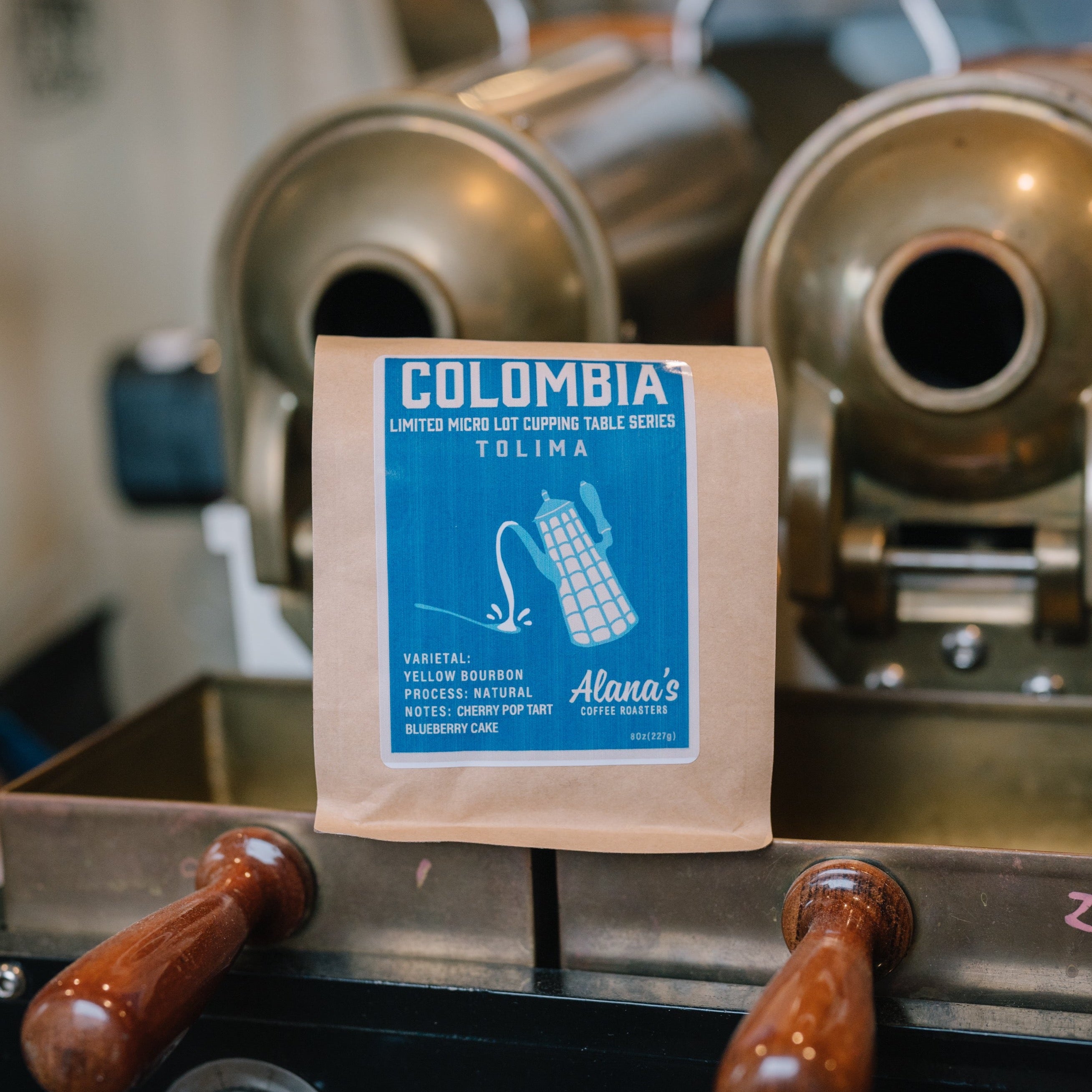 An Alana's Coffee Roasters' Tolima Colombian coffee bag staged in front of a coffee grinder. The label shows a white pitcher on a bright blue backdrop.