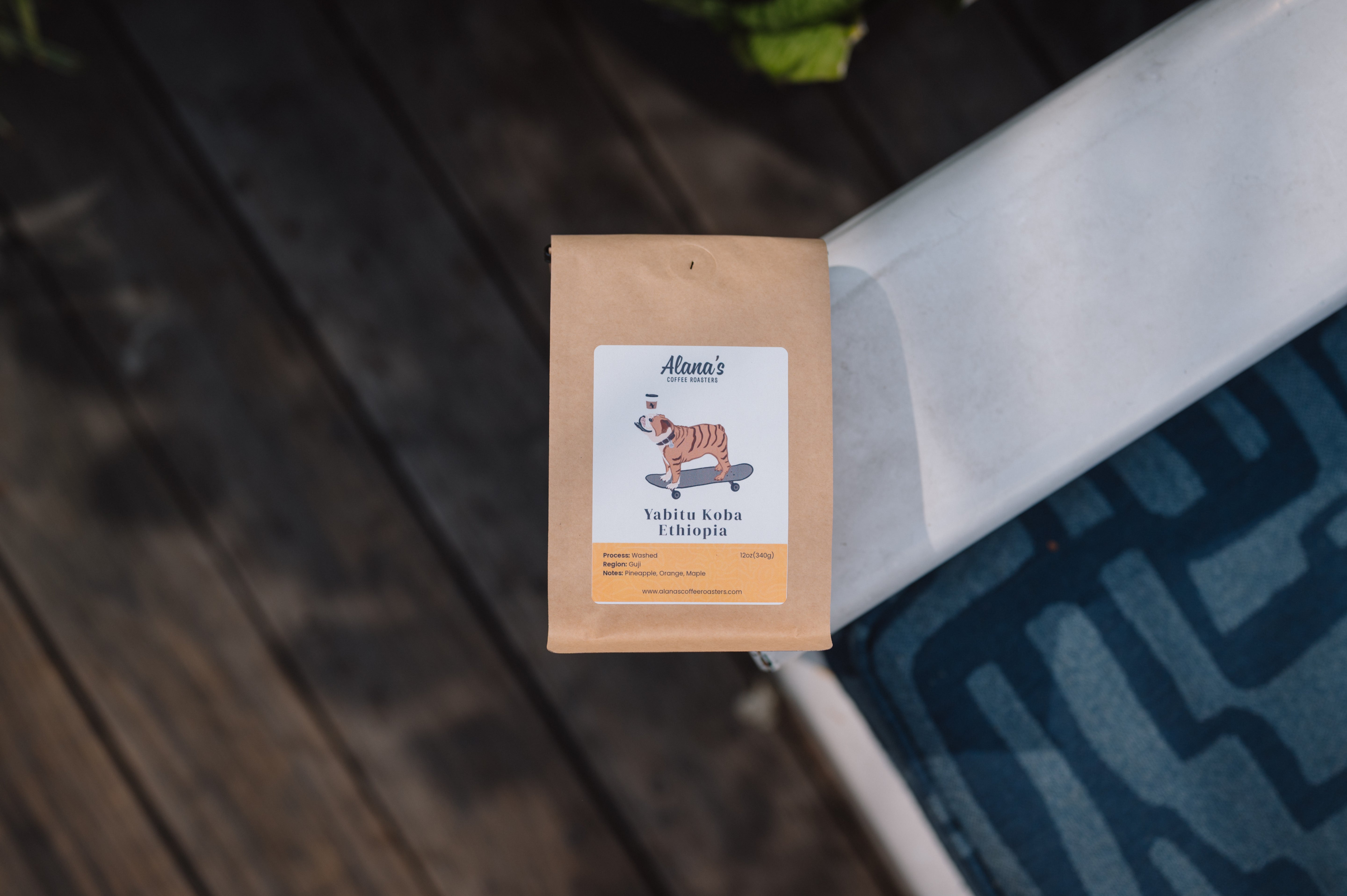 A kraft paper bag of Yabini Kola (Ethiopian) coffee by Anna's Coffee Roasters, resting on a dark wooden deck next to a white railing and a blue patterned rug.