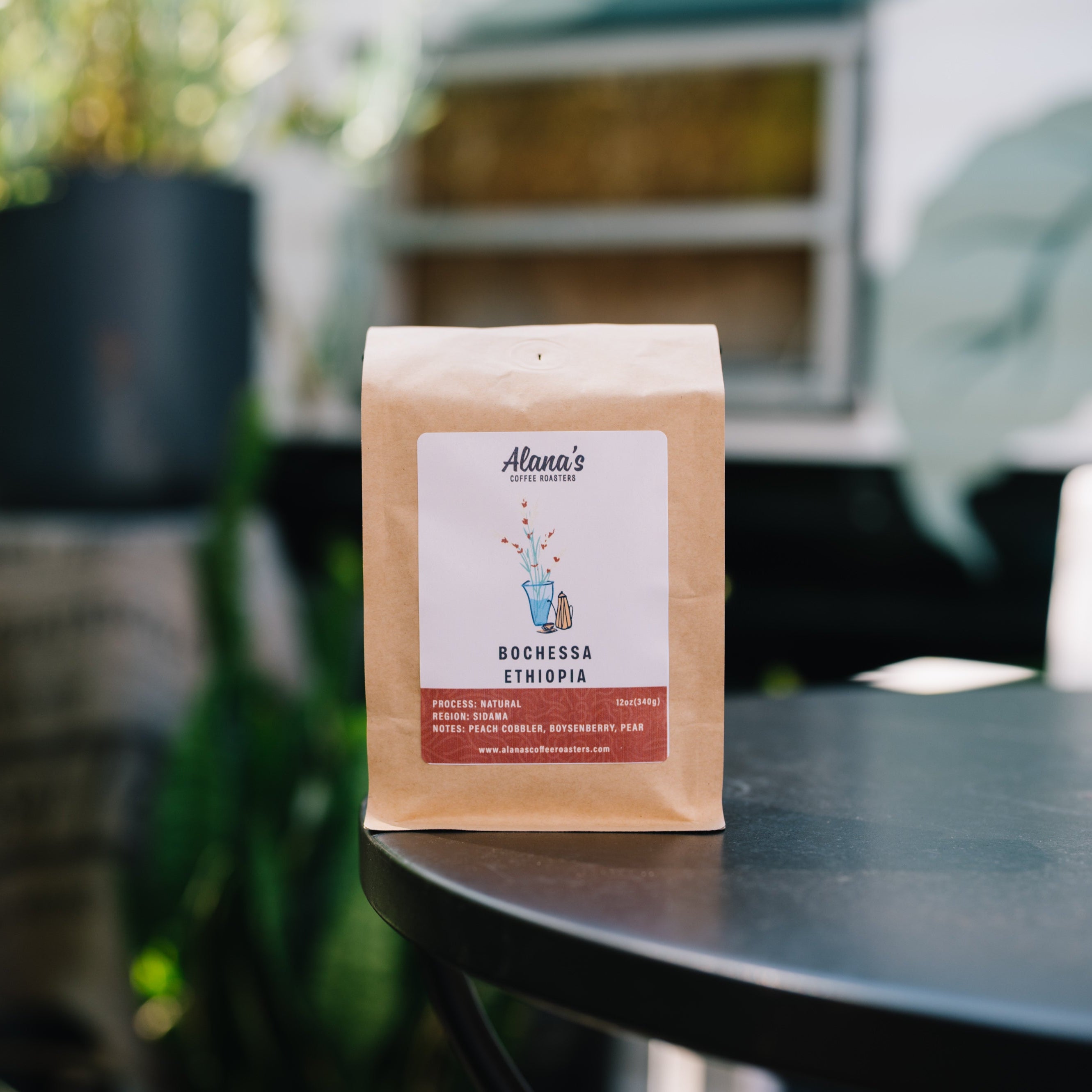 A bag of Aloma's Coffee Roasters Ethiopia coffee beans stands on a black table in a softly blurred outdoor patio setting.