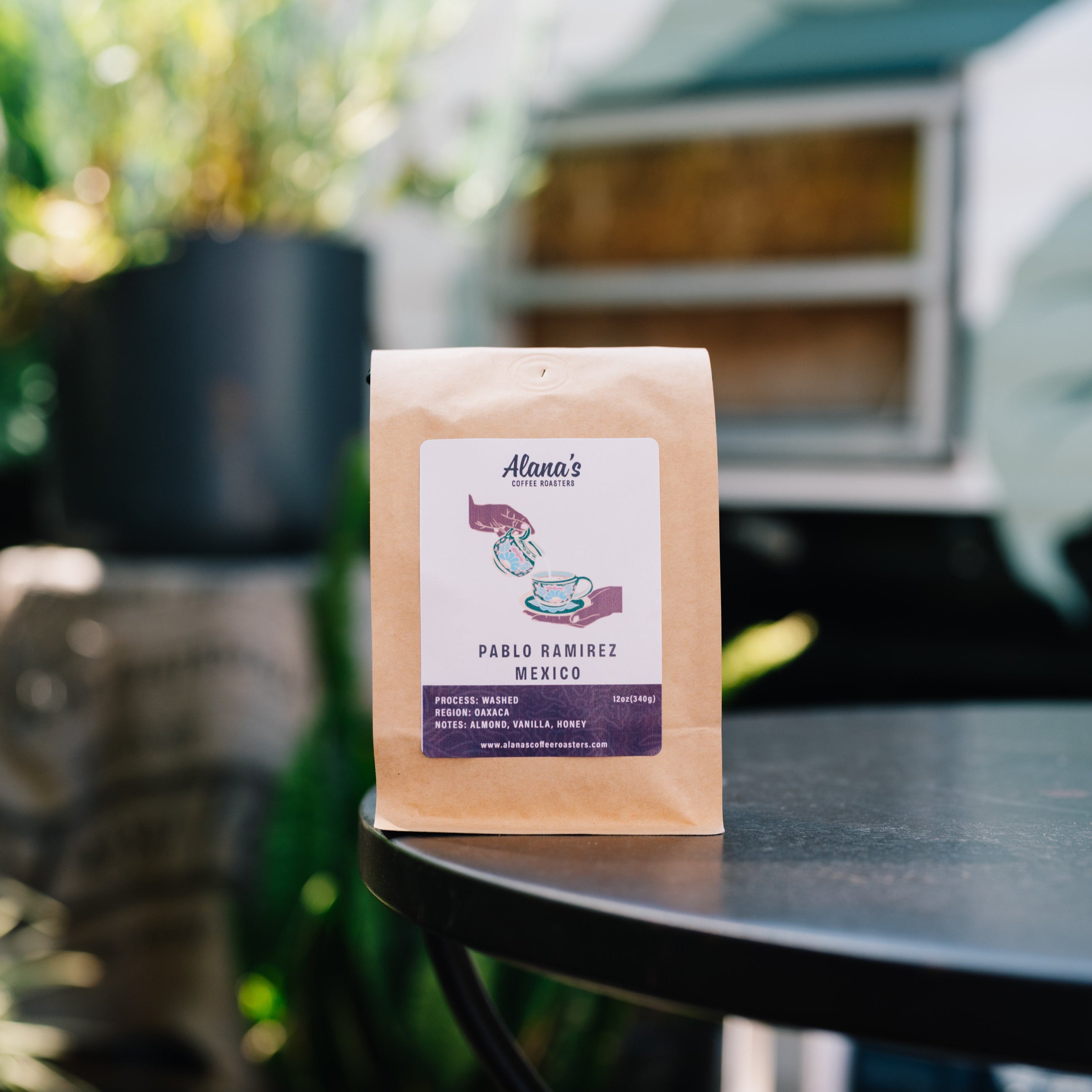 A stand-up kraft paper coffee bag labeled 'Alma's Coffee Roasters' and 'Pablo Ramirez México' sits on a dark round table in a softly blurred outdoor patio setting with green plants in the background.
